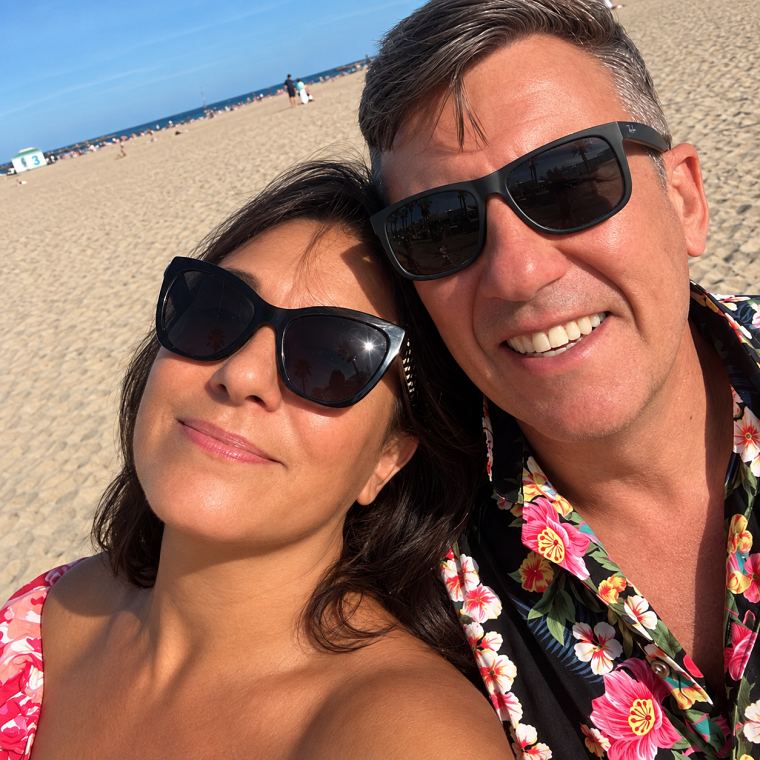 Louisa and Mark, smiling in sunglasses on a beach.